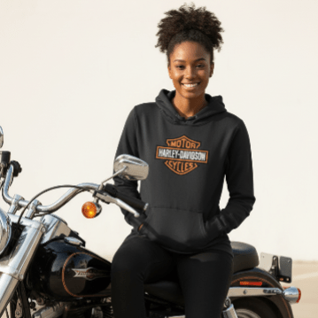 Harley-Davidson Womens Hoodies and black pants sits on a black cruiser motorcycle against a plain light background.