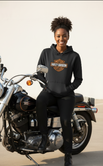 A woman wearing a black Harley-Davidson hoodie and black pants sits on a black cruiser motorcycle against a plain light background.