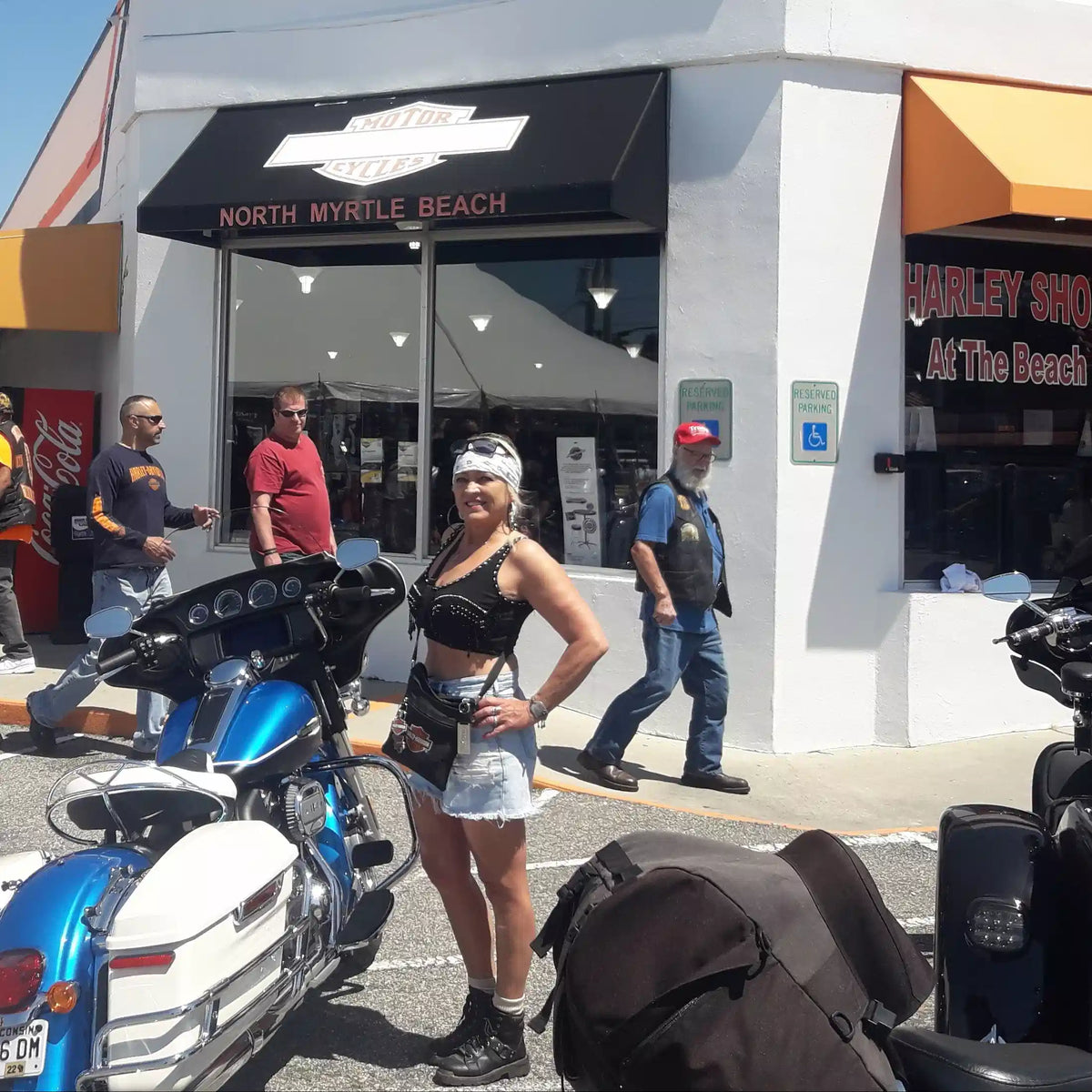 North Myrtle Beach Harley-Davidson, wearing a black crop top, denim skirt, and boots beside a blue and white motorcycle.